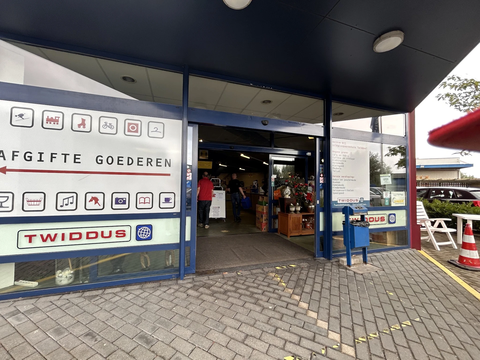 Gevel van kringloopwinkel Twiddus met blauwe pui, openbare deur en winkelraam met pictogrammen van aangeboden categorieën