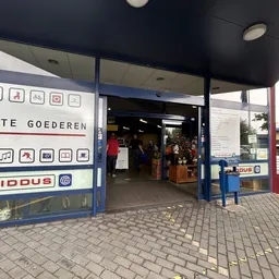Gevel van kringloopwinkel Twiddus met blauwe pui, openbare deur en winkelraam met pictogrammen van aangeboden categorieën
