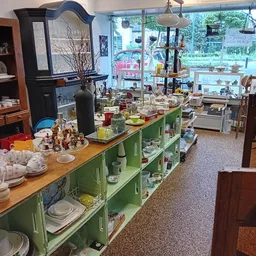 Winkelbinnenkant met groene kasten vol serviesgoed, houten tafel met decoratieve items en grote ramen met uitzicht op straat