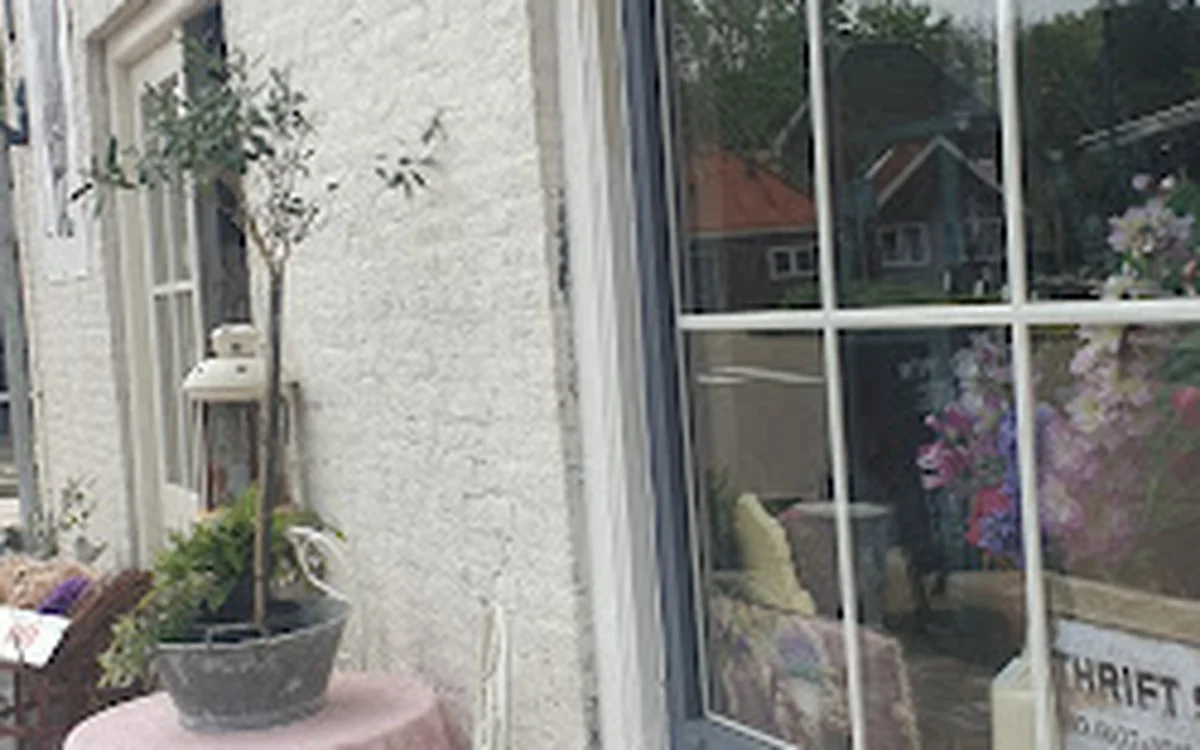 Witte gevel van kringloopwinkel met raam en ingangsdeur, potplanten op tafel buiten