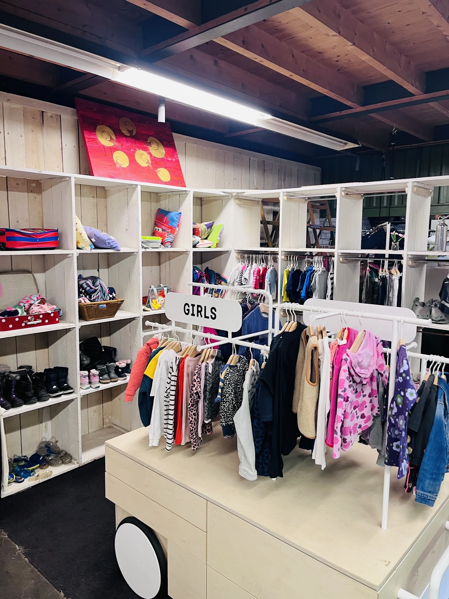Witte stellingen met tweedehands kinderkleding, schoenen en accessoires in een kringloopwinkel, met een 'GIRLS' bord in het m