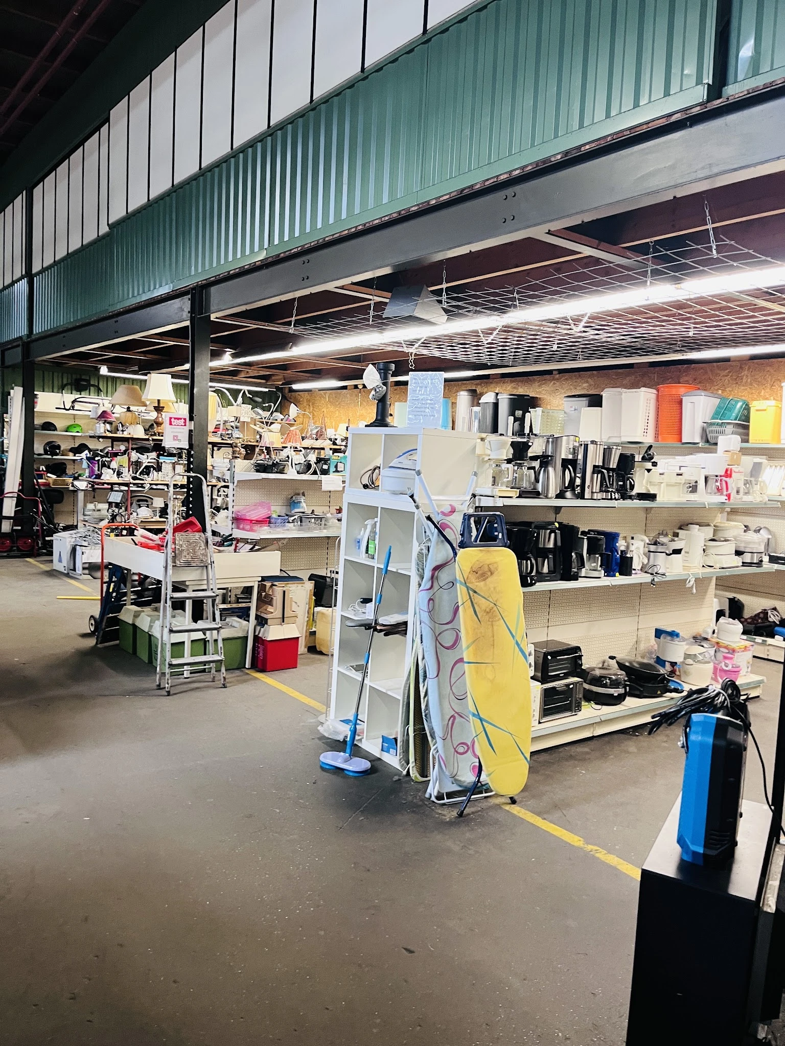 Winkelvloer van kringloopwinkel met stellingen, kasten en diverse spullen uitgestald, inclusief een geel surfboard