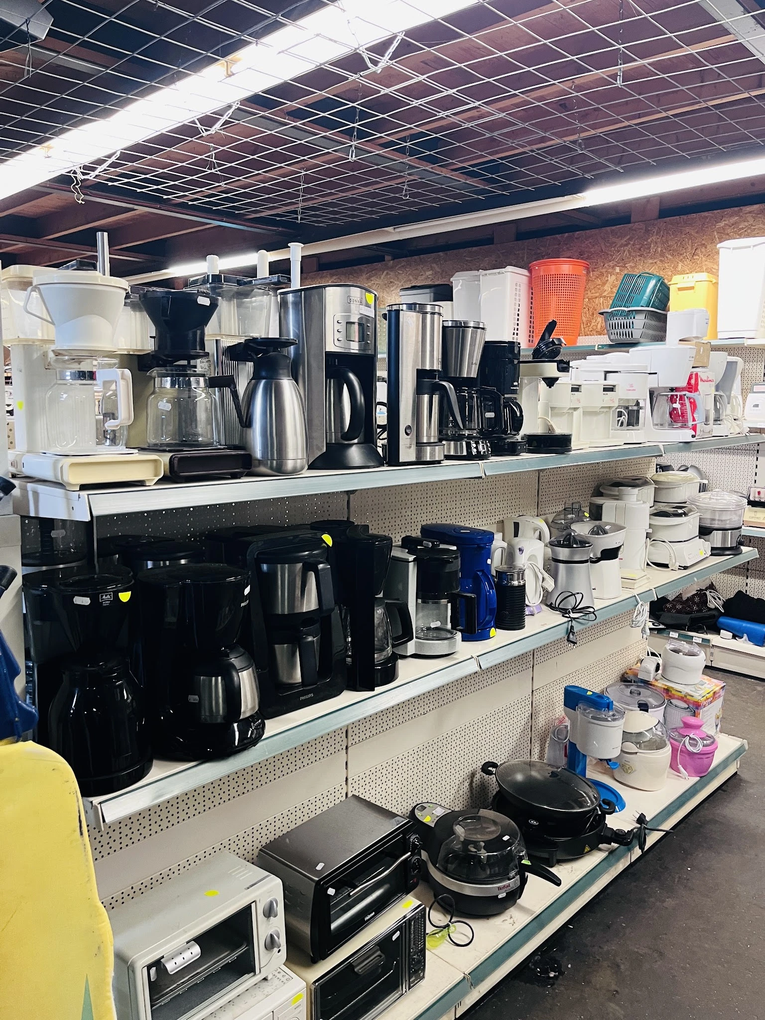 Uitgestalde keukenmachines en apparaten op schappen in een kringloopwinkel, waaronder koffiezetapparaten, waterkokers en mixe