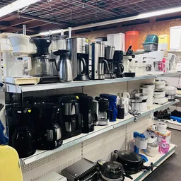 Uitgestalde keukenmachines en apparaten op schappen in een kringloopwinkel, waaronder koffiezetapparaten, waterkokers en mixe