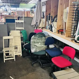 Diverse gebruikte meubels en stoelen in een kringloopwinkel, waaronder witte klapstoelen, bureaustoelen met rode en blauwe be