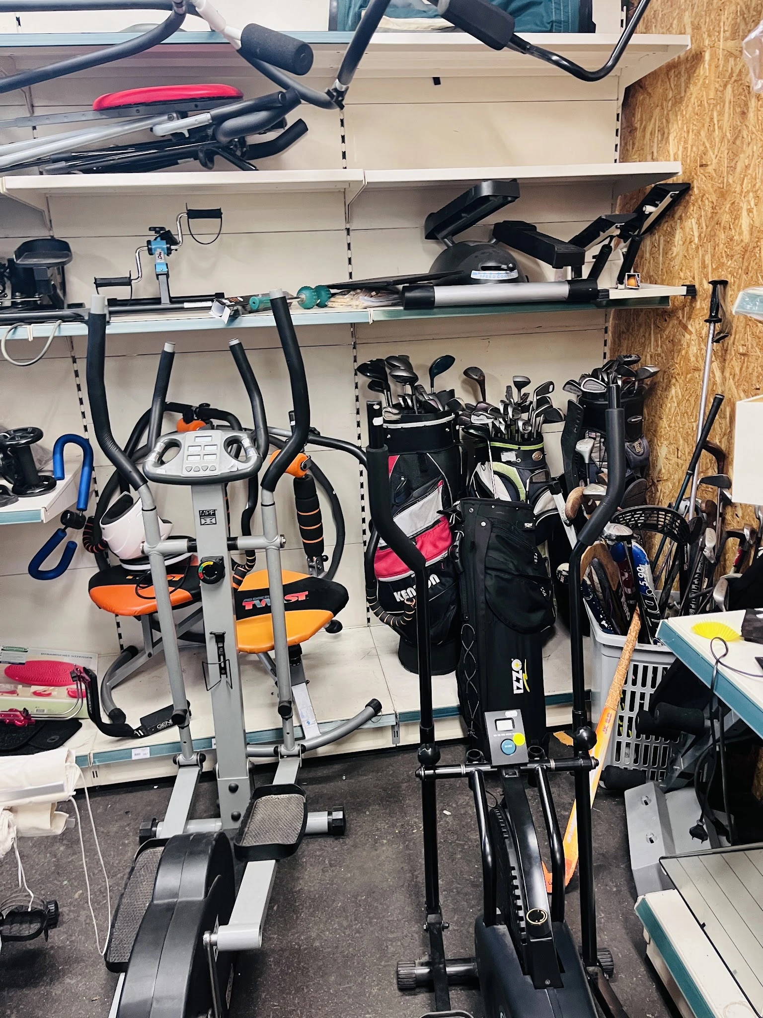 Stellingen met tweedehands sportartikelen en fietsonderdelen, waaronder hometrainers, golfclubs en fietsen