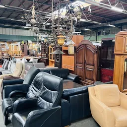 Uitgestalde meubels in kringloopwinkel: zwarte leren fauteuils, houten kasten en een crèmekleurige stoel op de winkelvloer