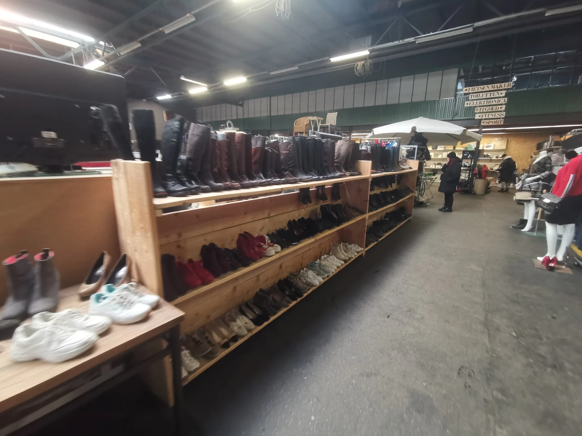 Winkelfloor met houten stellingen vol tweedehands kleding, neonverlichting, en klanten in de kringloopwinkel