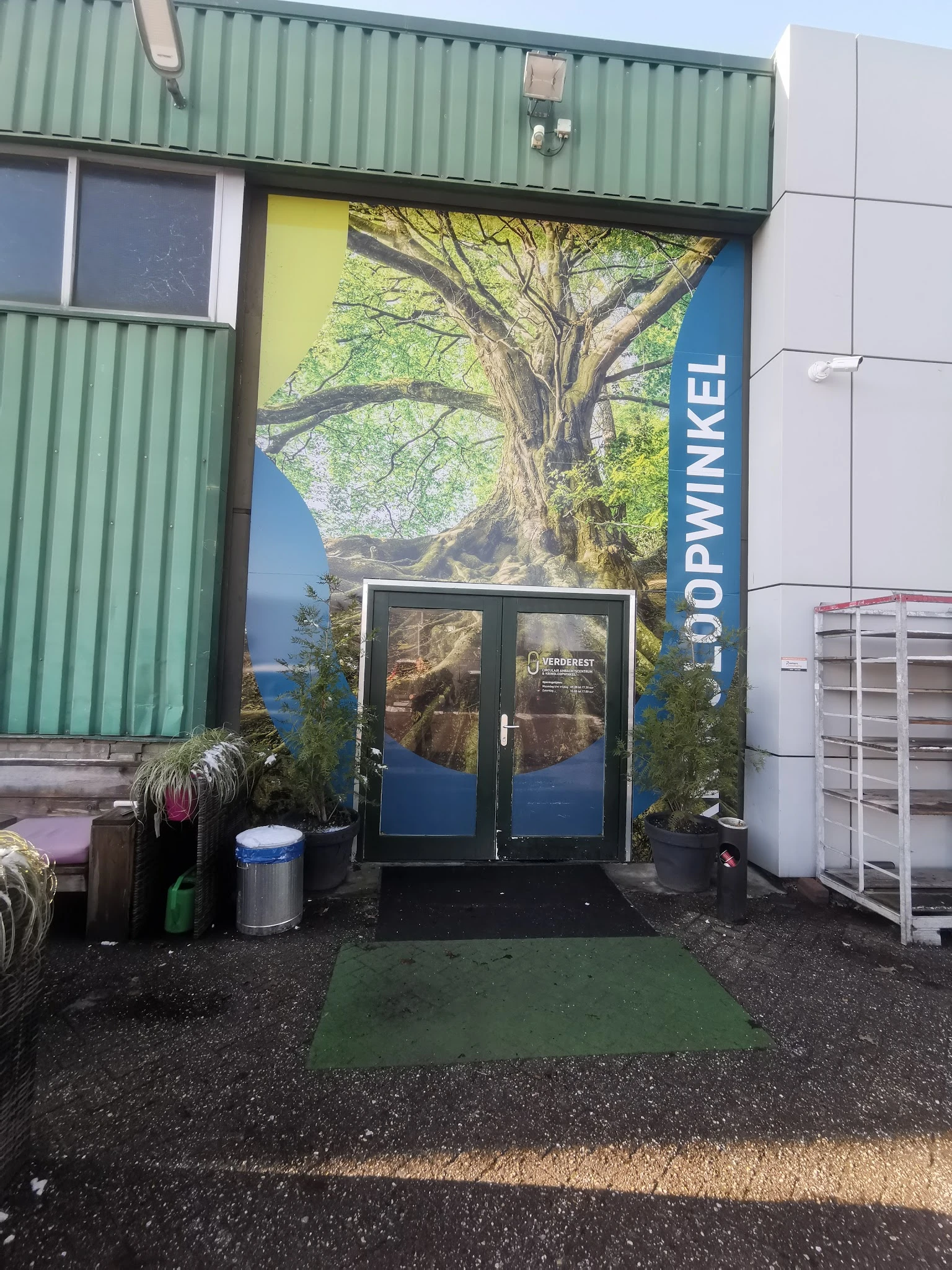 Ingang van kringloopwinkel met dubbele glazen deuren, grote boom-afbeelding op achtergrond, groene en witte zijwanden
