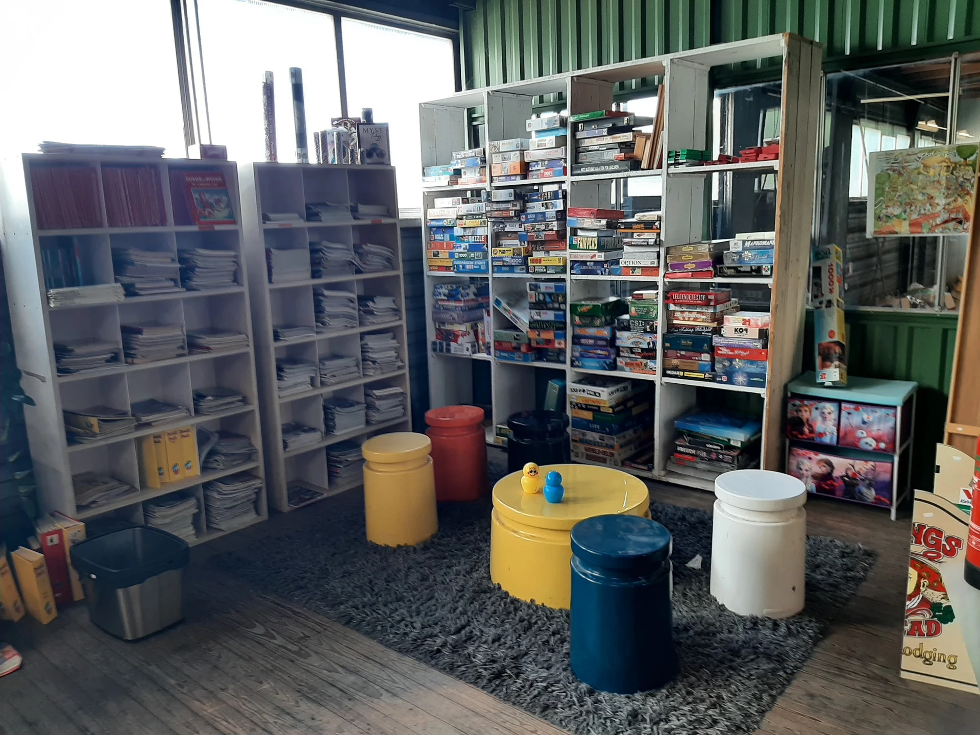 Winkelvloer met stellingen vol dvd's, boeken en spullen, enkele zitpoefjes en grijze vloerkleed