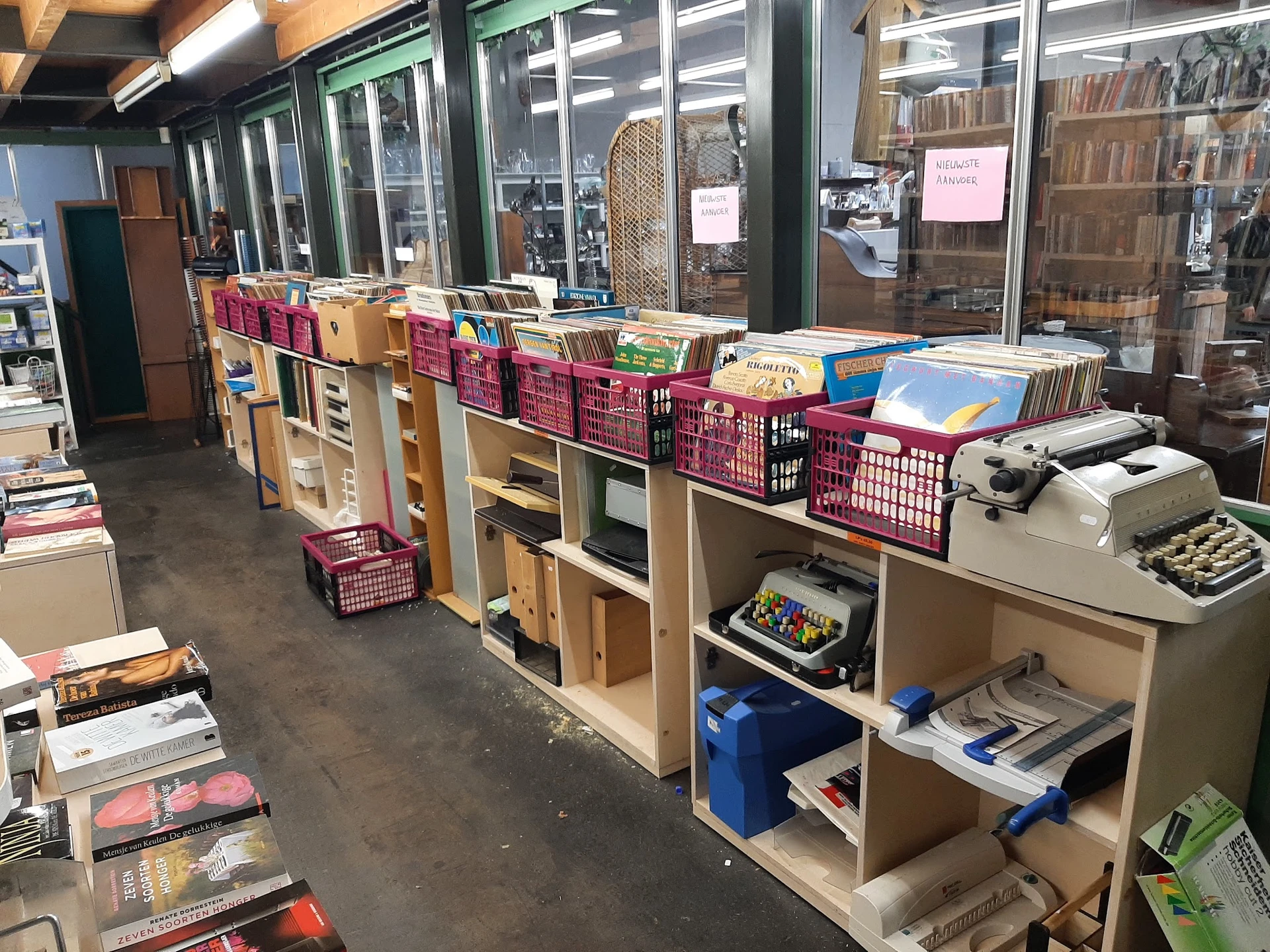 Winkelvloer met houten stellingen, roze kratjes, boeken en diversen spullen langs grote ramen