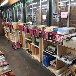 Winkelvloer met houten stellingen, roze kratjes, boeken en diversen spullen langs grote ramen