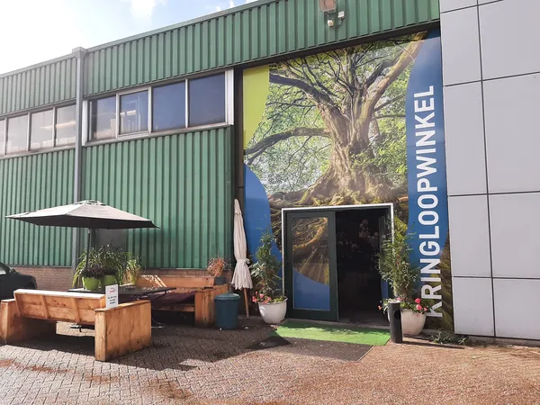 Voorkant kringloopwinkel met groene golfplaten gevel, grote boom-foto op ingang, zithoek met houten banken en parasol
