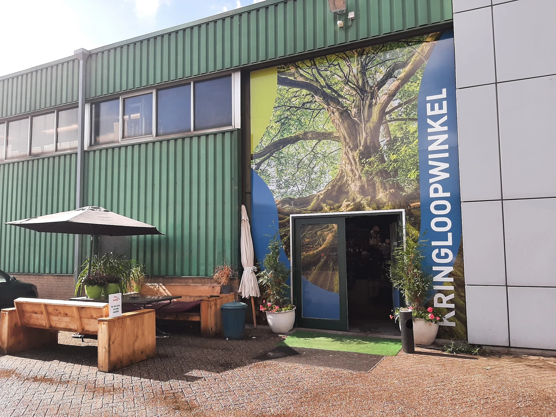 Voorkant kringloopwinkel met groene golfplaten gevel, grote boom-foto op ingang, zithoek met houten banken en parasol