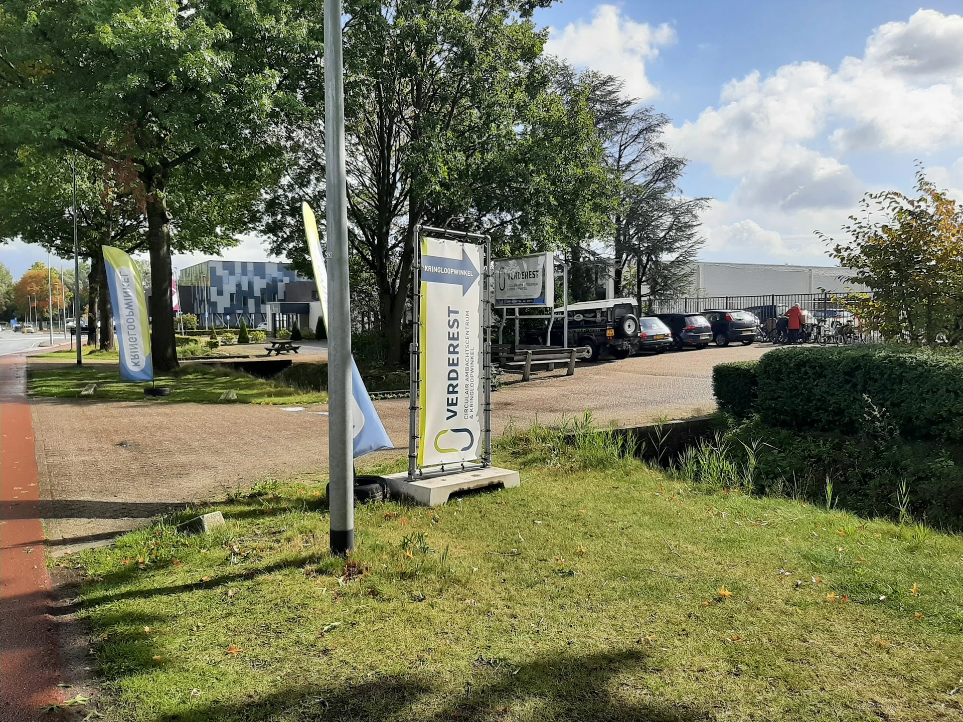 Exterieur van kringloopwinkel met parkeerplaats, bomen en bedrijfsgebouwen op achtergrond