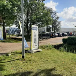 Exterieur van kringloopwinkel met parkeerplaats, bomen en bedrijfsgebouwen op achtergrond
