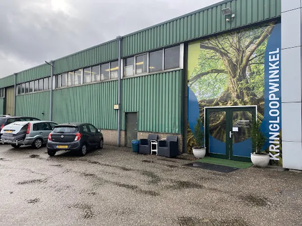 Groene gevel van kringloopwinkel met blauwe deur en grote boomafbeelding, enkele auto's op parkeerterrein