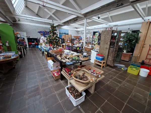 Overzicht van kringloopwinkel met tafels vol gebruiksartikelen, decoraties en spullen, hoog plafond met raamwerk