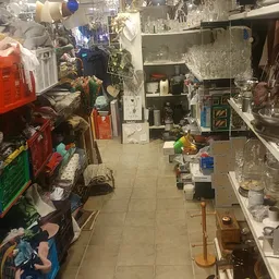 Rommelige winkelvloer met diverse spullen, dozen en artikelen links en rechts, stellingen met huishoudartikelen