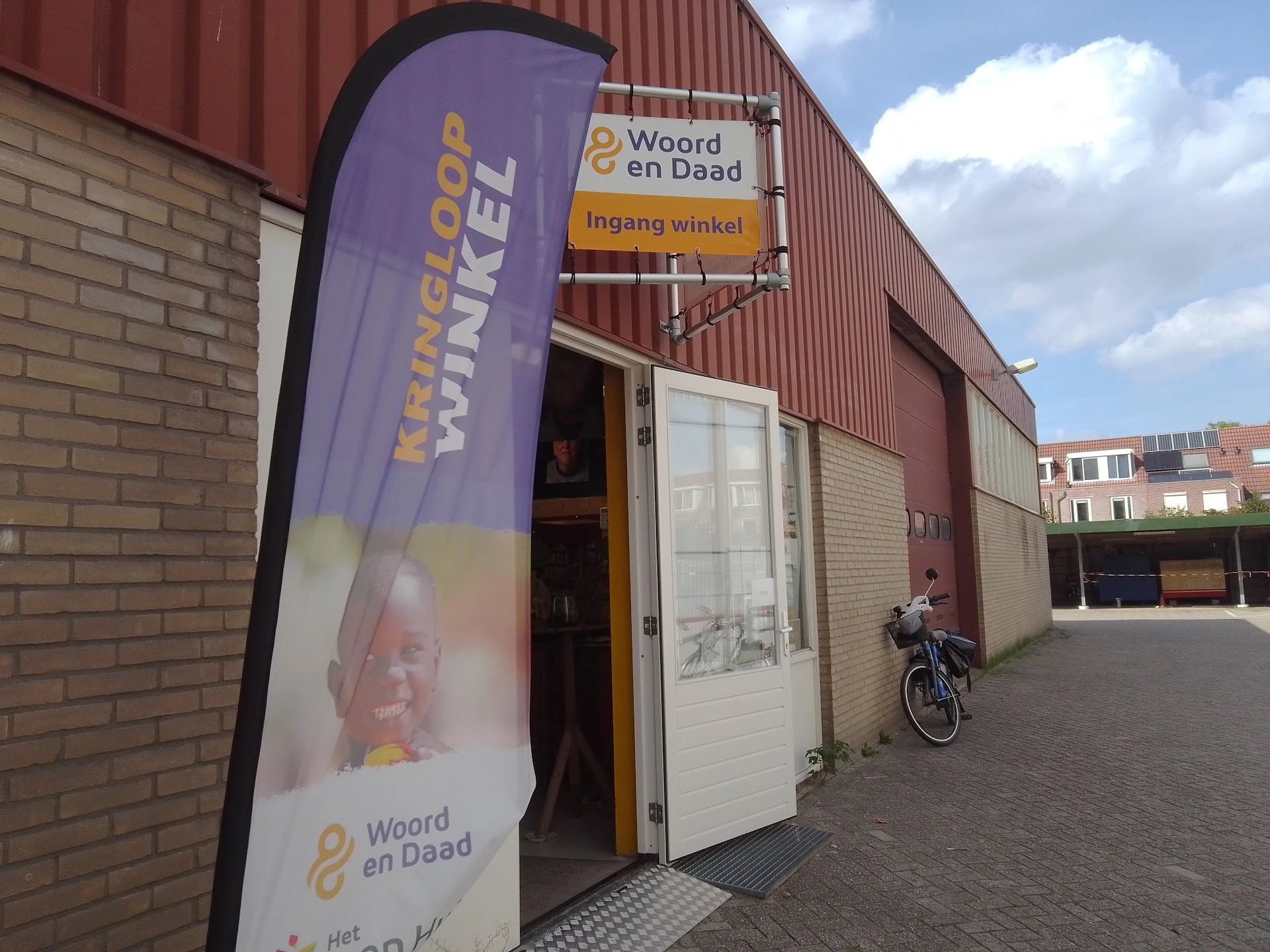 Voorgevel van kringloopwinkel met bakstenen muur, rode golfplaten, paarse reclamebanner en ingangsdeuren