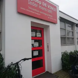 Voorkant van kringloopwinkel met rode deur, rode luifel met winkelnaam en wit gevel, fiets ervoor