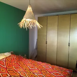 Slaapkamer met groen geschilderde wand, houten kledingkast en hanglamp van gevlochten materiaal boven bed met rood patroon