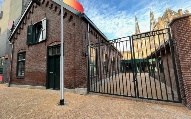 Bakstenen gevel van kringloopwinkel met zwarte deur, raam en zwart hek, op bakstenen plein