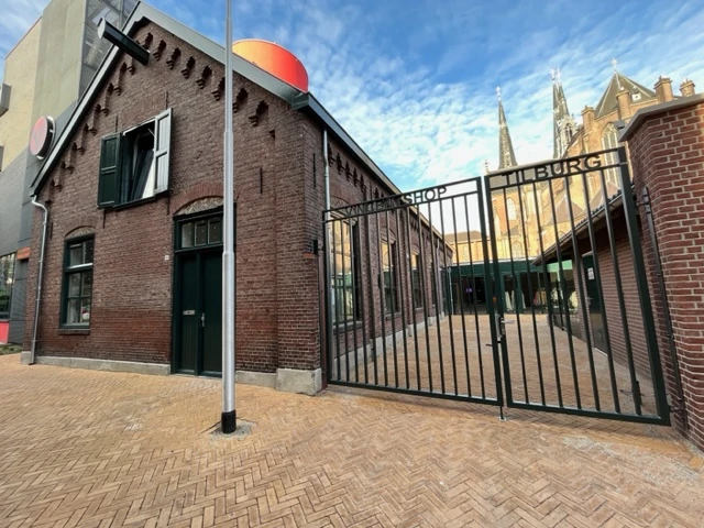 Bakstenen gevel van kringloopwinkel met zwarte deur, raam en zwart hek, op bakstenen plein