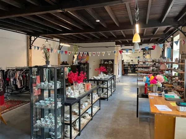 Ruim kringloopwinkelinterieur met kledingrekken links, glazen vitrines in het midden met decoratieve items, en diverse goeder