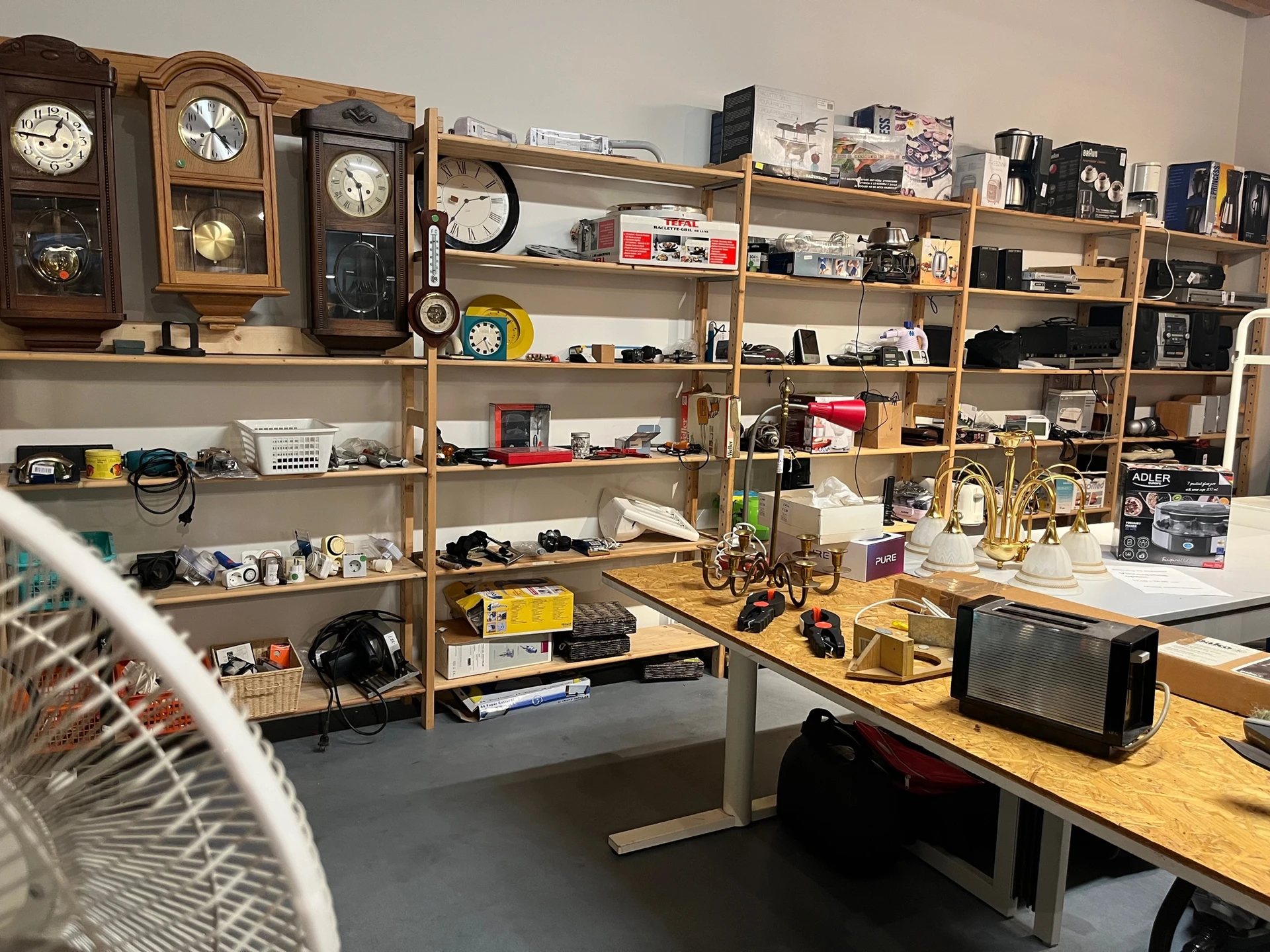 Werkruimte met houten stellingen vol elektronicaartikelen, klokken en gadgets, met werkbank en zwart televisietoestel.