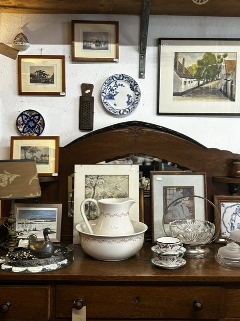 Uitgestalde vintage servies, decoratieve borden, beeldjes en fotolijsten op een donker houten meubel