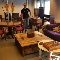 Winkelvloer met meubels: ronde tafel met stoelen, houten salontafel, paarse bank en diverse kasten. Een medewerker staat in h
