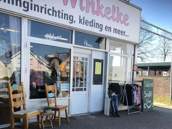 Gevel van kringloopwinkel met witte pui, ramen met etalage en ingang. Houten stoelen en kledingrek op het terras.
