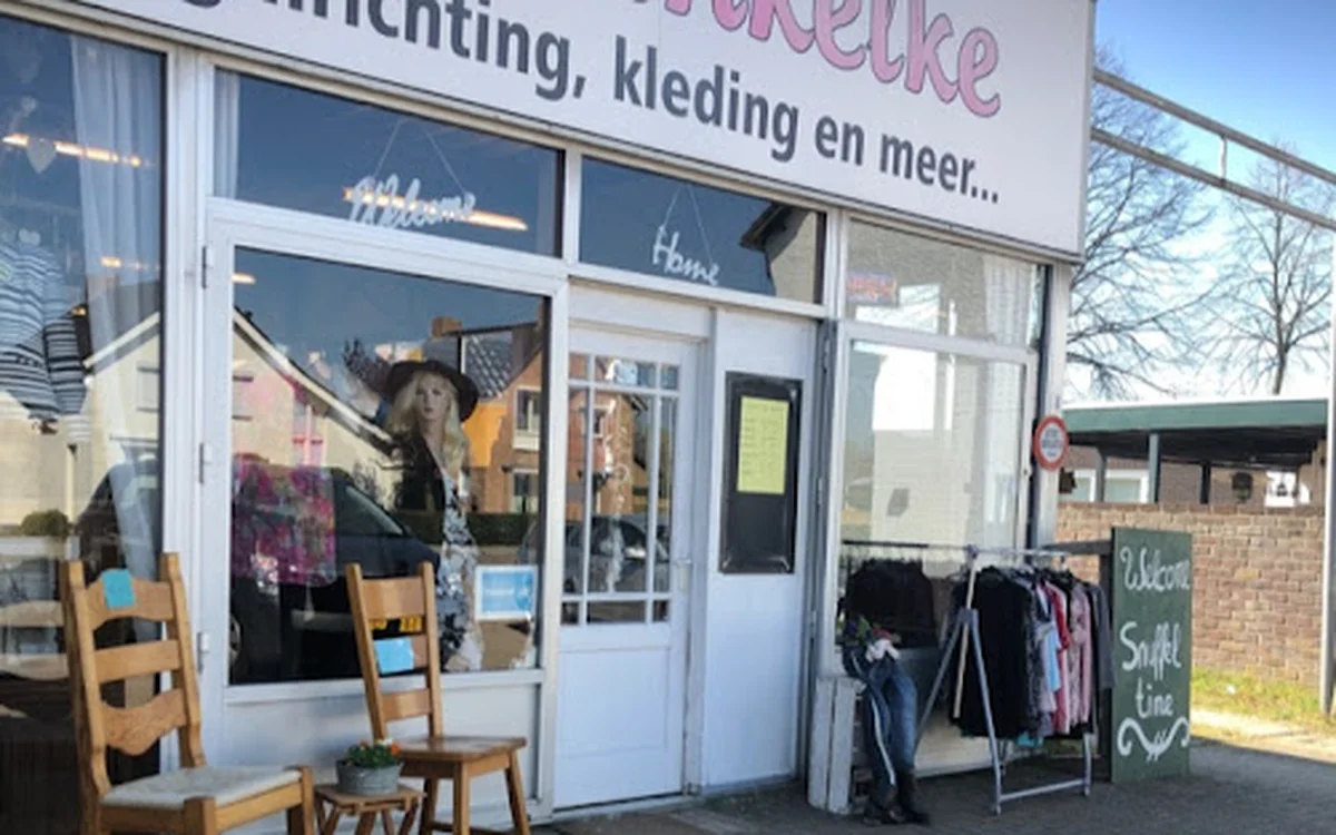 Gevel van kringloopwinkel met witte pui, ramen met etalage en ingang. Houten stoelen en kledingrek op het terras.