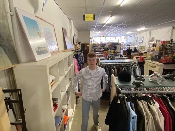 Mannelijk persoon staat in kringloopwinkel tussen rekken met kleding en shelves met diverse spullen