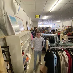 Mannelijk persoon staat in kringloopwinkel tussen rekken met kleding en shelves met diverse spullen