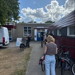 Voorgevel van kringloopwinkel met ingang, blauwe deur en enkele bezoekers buiten met fietsen