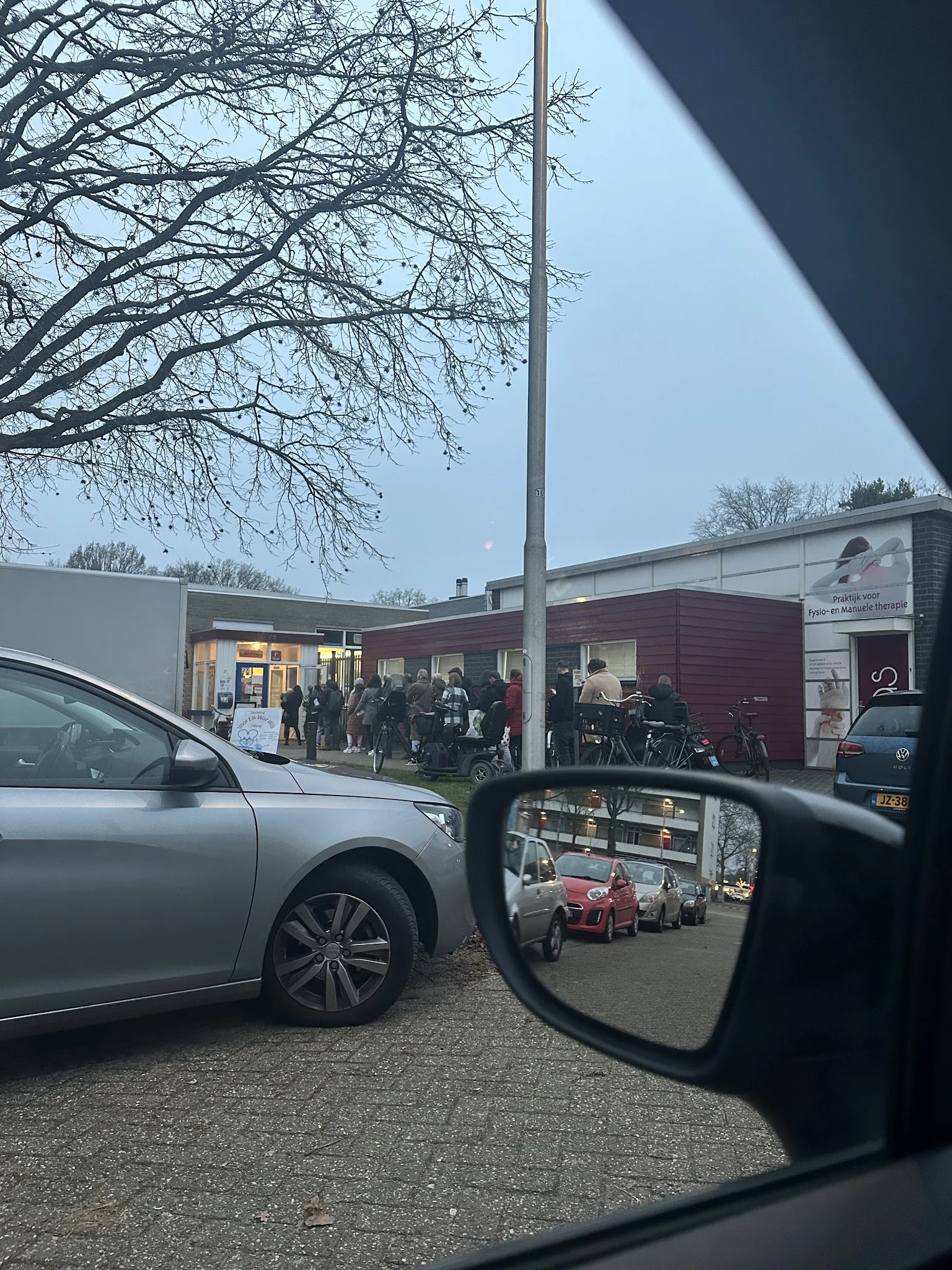 Voorgevel van kringloopwinkel met donkerrood en wit pand, bezoekers zichtbaar bij ingang, genomen vanaf parkeerplaats