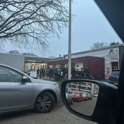 Voorgevel van kringloopwinkel met donkerrood en wit pand, bezoekers zichtbaar bij ingang, genomen vanaf parkeerplaats