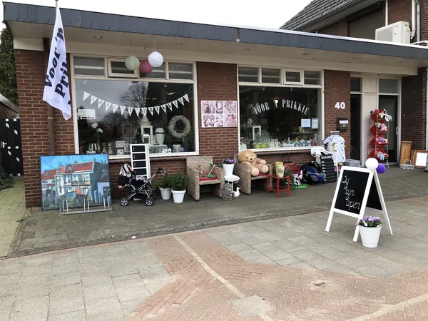Voorgevel van kringloopwinkel met etalage, uitgestalde spullen, witte vlaggen en reclameborden op het terras