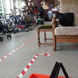 Grijze betonvloer met rood-wit markeringstape en houten stoelen, op de achtergrond fietsen en meubels