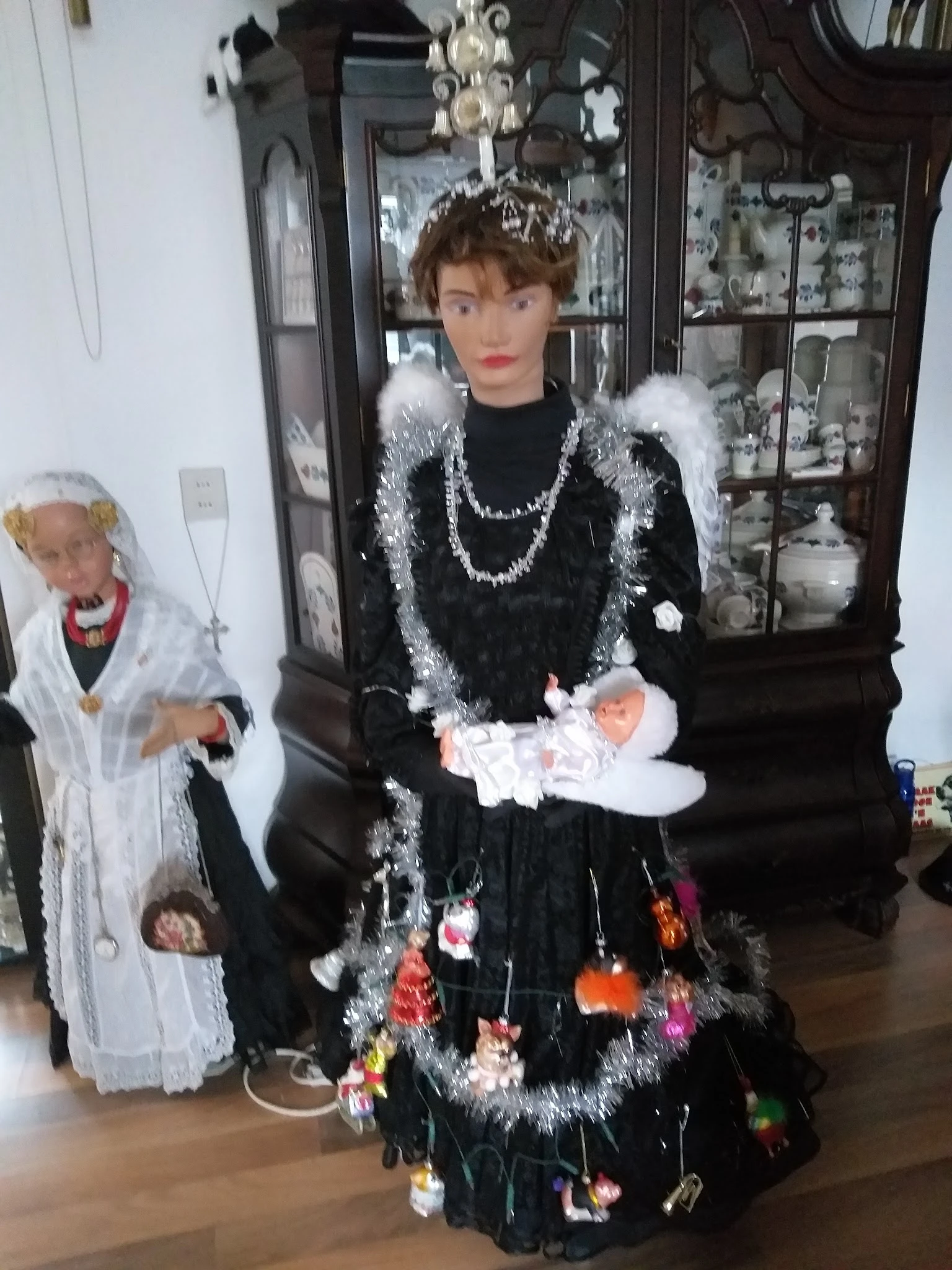 Etalagepop in zwarte jurk met witte bontafwerking en sieraden, achter vitrinekast met servies