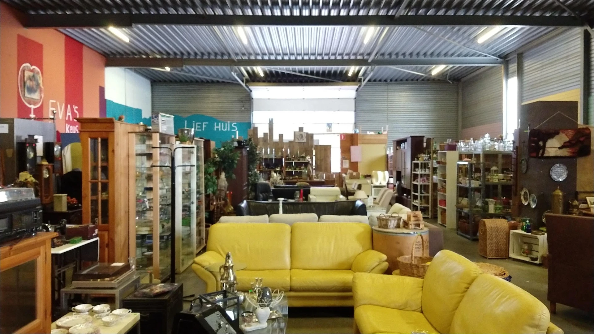 Winkelinterieur met gele banken en fauteuils, stellingen met huishoudartikelen en decoratie, industrieel plafond met daglicht