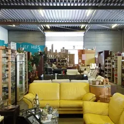 Winkelinterieur met gele banken en fauteuils, stellingen met huishoudartikelen en decoratie, industrieel plafond met daglicht