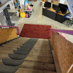 Trap naar beneden met grijze traptreden en houten zijwanden, rode mat in de hal en roze/paarse wanden