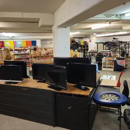 Computerworkstations met drie monitoren en bureaustoel in een kringloopwinkel, met stellingen met spullen op de achtergrond