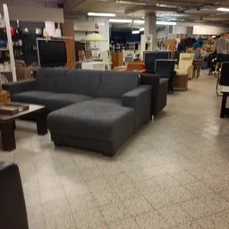 Grijze hoekbank en houten tafels in de winkel, met diverse meubels en spullen op de achtergrond