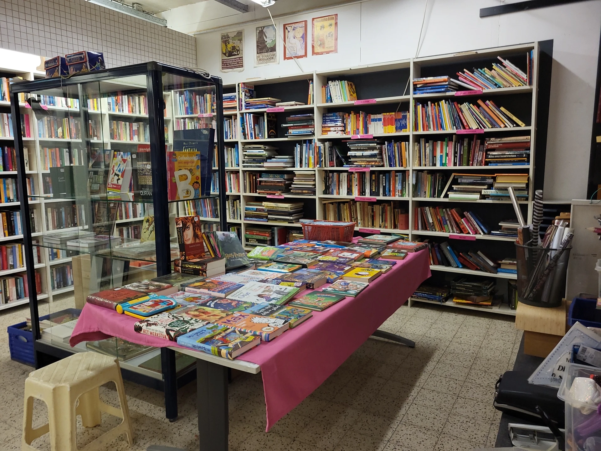 Uitgestalde boeken en strips op een tafel met roze kleed in een boekenkringloop, met boekenkasten op de achtergrond