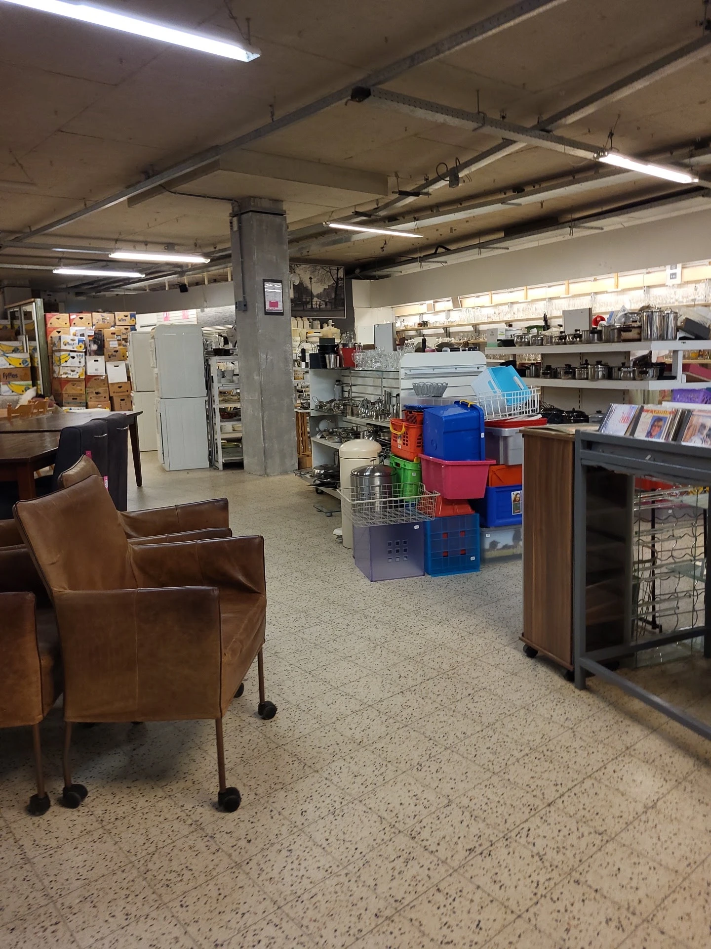 Winkelvloer van kringloopwinkel met stellingen, huisraad, een bruine leunstoel en diverse tweedehands artikelen