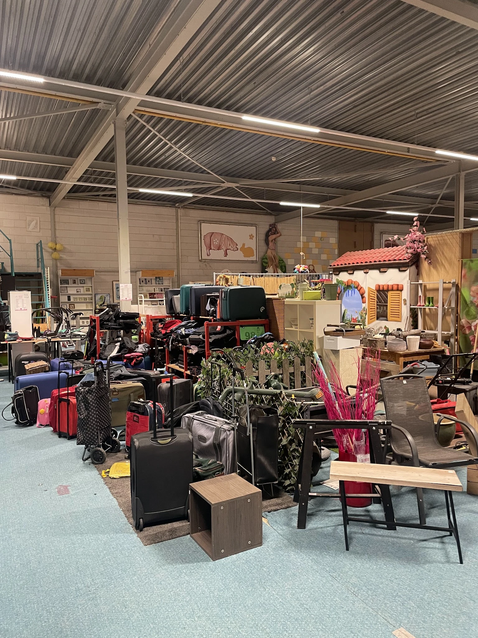 Overzicht van de kringloopwinkel met kledingrekken, koffers, huishoudspullen en meubels verspreid over de vloer